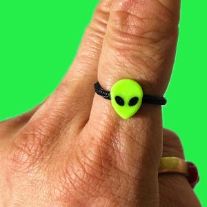 May include: A neon green alien ring with black eyes and a black band. The ring is worn on a finger against a bright green background. The ring is a fun, quirky accessory.