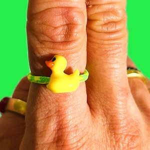 May include: A yellow rubber duck ring with a green and yellow band. The duck has black eyes and an orange beak. Other rings are visible in the background.