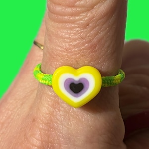 May include: A handmade ring featuring a yellow heart-shaped bead with purple and black accents. The ring band is a neon green and yellow braided cord. The ring is worn on a finger against a bright green background.
