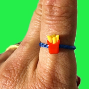 May include: A novelty ring featuring a miniature red carton of french fries with yellow fries. The ring band is a blue cord. The ring is on a finger against a green background.