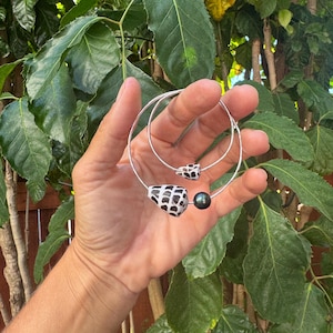 Puede incluir: Una pulsera de aro hecha de alambre plateado que presenta dos cuentas en forma de cono con un patrón geométrico en blanco y negro, junto con una cuenta esférica de color gris oscuro. La pulsera tiene una estructura de metal circular.