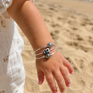 May include: A set of three silver-toned bangles worn on a wrist. Two bangles feature a single dark grey spherical bead, while the center bangle features a small, white, triangular-shaped charm with black spots, resembling a cow print pattern.