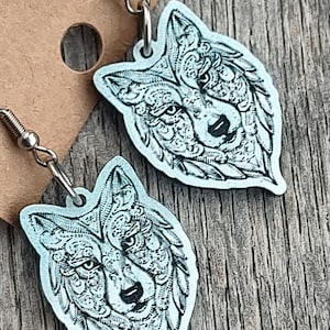May include: A pair of light blue wolf-shaped earrings with intricate etched details. The earrings hang from silver-colored hooks and are displayed on a brown cardboard backing, set against a wooden surface.