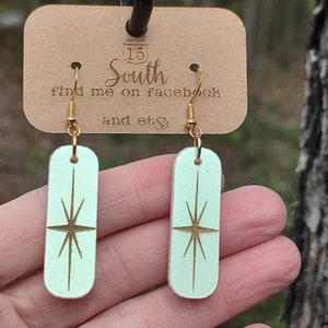 May include: A pair of light green wooden earrings with a gold starburst design. The earrings are on a brown card with the text "15 South find me on facebook and etsy".