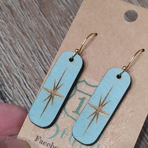 May include: A pair of light blue wooden earrings with a gold starburst design and gold hoops. The earrings are displayed on a brown card with the text "Facebook"