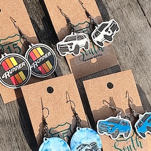 May include: Earrings with car designs on brown cards. One pair features a circular design with the word "Runner" and a striped pattern in red, orange, yellow, and black. Other pairs have car silhouettes in white and blue.