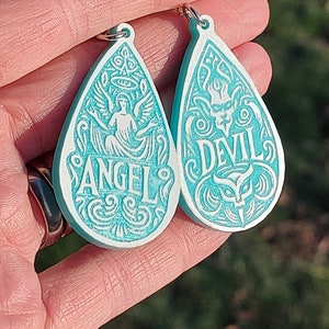 Op de afbeelding: Twee tealblauwe druppelvormige hangers met witte ontwerpen. De ene hanger zegt "ANGEL" en heeft een engel met vleugels. De andere hanger zegt "DEVIL" en heeft een duivel met hoorns.