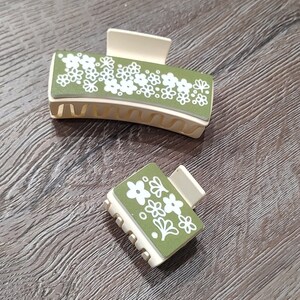 Pode incluir: Dois prendedores de cabelo com um design floral. O prendedor maior é bege com um padrão floral verde. O prendedor menor é bege com um padrão floral verde.