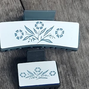 May include: Two blue and white hair clips with a floral design. The larger clip is curved and the smaller clip is rectangular.
