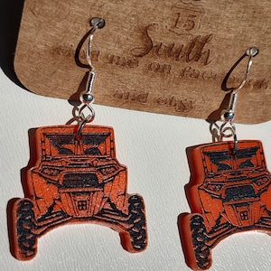 May include: Orange and black earrings shaped like a side-by-side off-road vehicle. The earrings are made of wood and have silver hooks.