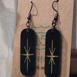 May include: A pair of black dangle earrings with gold starburst designs. The earrings are on a brown card with the text "15" and "look and"
