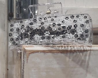 May include: A clear plastic hair clip with a paw print design. The clip is closed and sitting on a white surface.