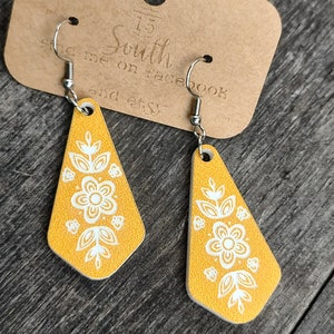 May include: A pair of yellow dangle earrings with a white floral design. The earrings are made of wood and have silver hooks.
