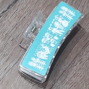 May include: A clear plastic hair clip with a blue and white design. The design features a white rooster and other white figures on a light blue background.