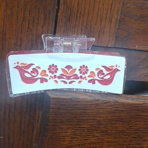 May include: A clear plastic hair clip with a red, white, and orange floral design. The design features two birds and flowers.