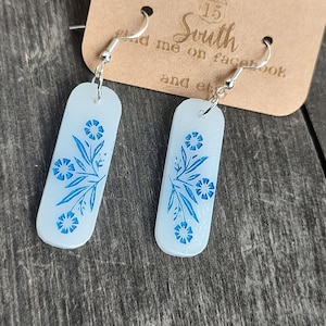 May include: A pair of rectangular earrings with a light blue background and a blue floral design. The earrings hang from silver hooks. A brown tag with the text "South" is in the background.