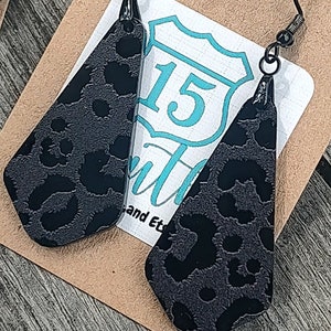 May include: A pair of black dangle earrings with a leopard print design. The earrings are made of a textured material and have a silver hook.