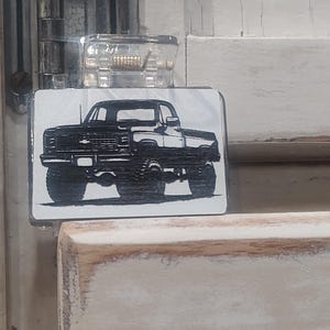 May include: A black and white graphic of a pickup truck on a rectangular metal sign. The truck is a classic model, with a detailed depiction of its front, side, and wheels. The sign is mounted on a light-colored wooden surface.