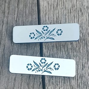 May include: Two rectangular, light blue decorative pieces with a floral design in dark blue. The floral design includes three small flowers and leaves. The pieces are on a wooden surface.