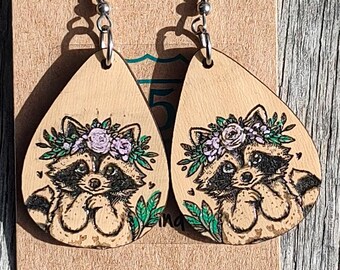 May include: Wooden teardrop earrings featuring a raccoon design. The raccoon is wearing a floral crown with purple flowers and green leaves. The earrings hang from silver-colored hooks.