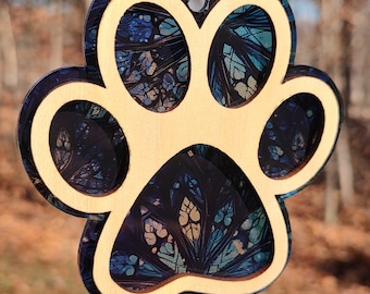 May include: A wooden paw print ornament with a blue and white abstract design. The ornament is hanging from a silver ribbon.