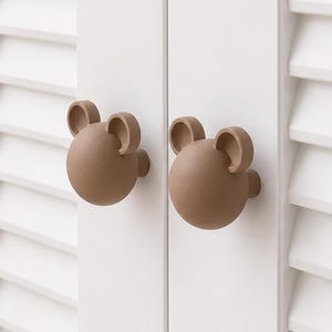 May include: Two light brown, spherical cabinet knobs shaped like bear heads with small rounded ears. These matte-finished knobs are mounted onto the vertical panels of a white louvered door, serving as functional handles for opening the cabinet.