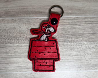 May include: A red Snoopy keychain featuring Snoopy on top of his doghouse. The keychain has a black outline and a silver key ring. The doghouse has black stitching and small black dots. The Snoopy figure is white with black details.