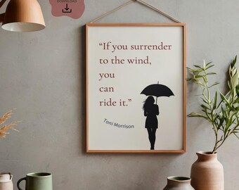 May include: A framed print with the quote "If you surrender to the wind, you can ride it." by Toni Morrison. The print features a silhouette of a person with an umbrella. The frame is brown, and the background is a neutral color.