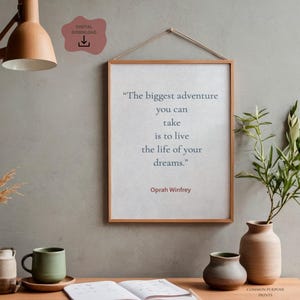 May include: A framed print with the quote "The biggest adventure you can take is to live the life of your dreams" by Oprah Winfrey. The print is in a wooden frame and hangs on a gray wall. A wooden desk holds a mug, vases, and an open book.