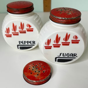 May include: Vintage white ceramic salt and pepper shakers with red lids and black lettering. The shakers have a design of red potted plants with a black outline. The salt shaker says "Sugar" and the pepper shaker says "Pepper".