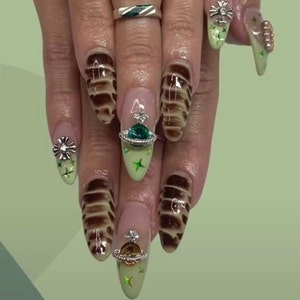 Puede incluir: Primer plano de una mano con uñas largas y de forma almendrada. Las uñas están pintadas con una base verde claro y tienen una variedad de diseños, que incluyen remolinos marrones, purpurina verde y abalorios plateados. Una uña presenta un abalorio plateado con un orbe verde y estrellas.