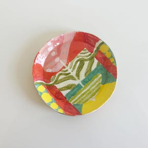 May include: A round ceramic dish featuring a hand-painted abstract design with bold sections of red, bright yellow, and green. The surface displays wavy green lines, solid color blocks, and small yellow dots, all finished with a glossy, reflective glaze.