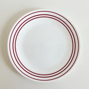 May include: A round white ceramic dinner plate featuring a decorative border consisting of two concentric rings in a deep red color. Each ring is composed of a repeating pattern of small, connected dots, creating a textured visual effect.