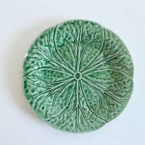 May include: A round ceramic plate featuring a raised, textured surface that mimics the appearance of a green cabbage leaf. The design includes detailed veins radiating from a central point, finished with a glossy, light to dark green glaze.