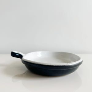 May include: A small, round ceramic dish with a short handle, featuring a smooth white interior and a matte black exterior. The rim shows a subtle transition where the white glaze meets the dark outer finish, creating a simple, clean aesthetic.