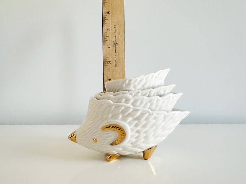 May include: A white ceramic hedgehog-shaped measuring cup set with gold accents. The set includes four nesting cups, each shaped like a leaf. The hedgehog has gold feet, a gold eye, and a gold crescent moon design.