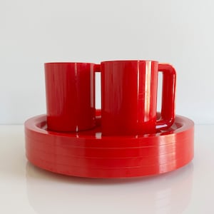 May include: A set of four red plastic plates and two red plastic mugs. The plates are stacked on top of each other and the mugs are sitting on top of the plates.