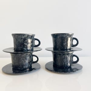 May include: Four black ceramic cups and saucers. The cups and saucers have a marbled appearance with a dark gray and white pattern. The cups have a rounded handle and sit atop matching saucers. The set is displayed on a white surface.