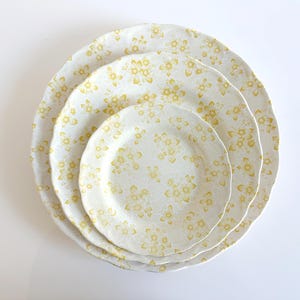 May include: A set of three white ceramic plates with a yellow floral pattern. The plates are stacked, with a large plate at the bottom, a medium plate in the middle, and a small plate on top. The edges of the plates have a wavy design.