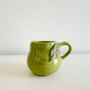May include: A round, lime green ceramic mug with a glossy finish. The mug features a sculpted, textured leaf detail attached to the side near the handle. The body of the mug has a bulbous, organic shape with a smooth, reflective surface throughout.