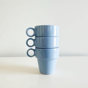 May include: A stack of three light blue ceramic mugs with ring handles. The mugs are cylindrical with a slightly flared base and are stacked on top of each other. The background is a plain white surface.