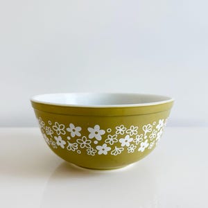 May include: A vintage olive green mixing bowl with a white floral pattern. The bowl has a white interior and a rounded shape. This retro kitchenware item is perfect for baking and food preparation.