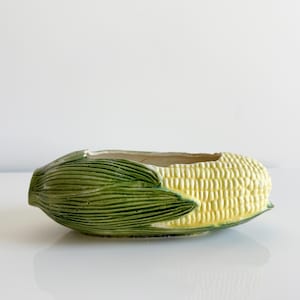 May include: A ceramic corn-shaped planter. The planter features a yellow corn cob design with green husks on each end. The planter is a decorative item for home decor.