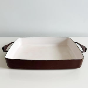 May include: A rectangular baking dish with a white interior and a dark brown exterior. The dish has two curved handles on either side. The baking dish is sitting on a white surface.