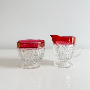 May include: A clear glass sugar bowl and creamer set with a ruby red rim. The glass has a diamond pattern. The sugar bowl has a lid with a silver rim. The creamer has a handle and spout.