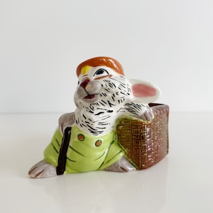 May include: A ceramic planter shaped like a rabbit wearing a brown hat and green overalls. The rabbit is holding a brown basket in its right hand. The planter is painted with white, brown, black, green, and pink details.