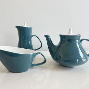 May include: A set of three teal ceramic pieces including a teapot, a creamer, and a pitcher. Each item features a smooth, glossy teal exterior with a contrasting white interior and a small, pointed white lid handle on the teapot and pitcher.