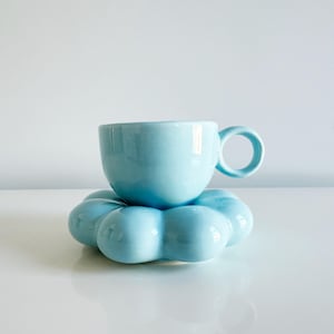 May include: A light blue ceramic teacup with a rounded handle sits atop a matching, flower-shaped saucer. The cup and saucer have a glossy finish and are set against a white background. This set is ideal for serving tea or coffee.