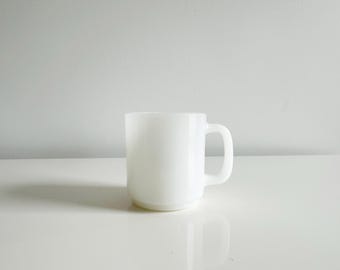 May include: A white ceramic mug with a rounded base and a curved handle. The mug is plain and has a smooth, opaque surface. The mug is sitting on a white surface.