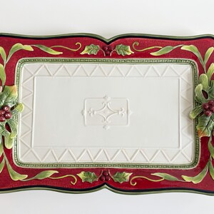 May include: A rectangular ceramic serving tray featuring a white center with a raised geometric pattern and a central embossed motif. The border is deep red with green holly leaves and red berries on the sides, finished with a dark green edge.
